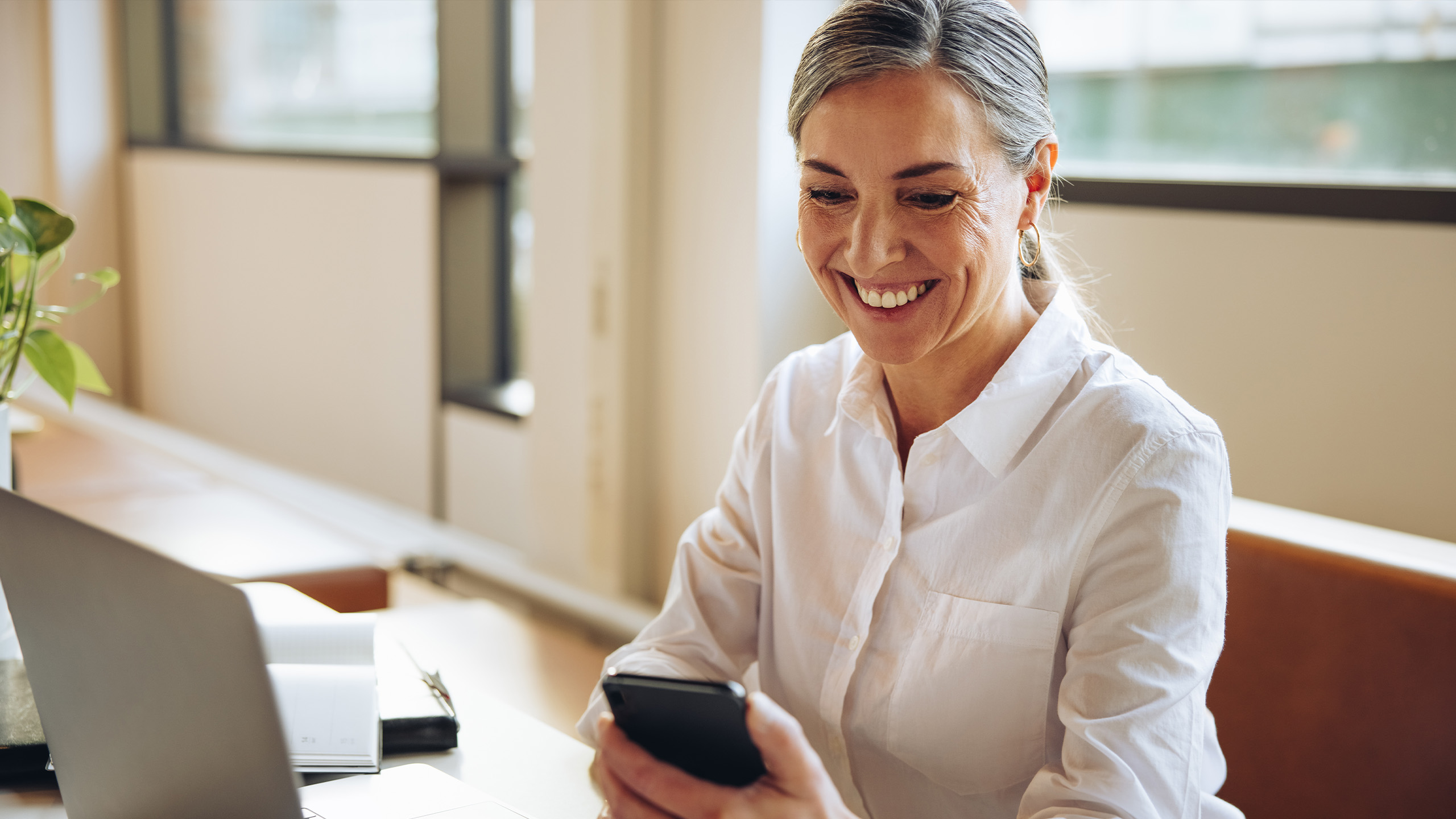 Smilende kvinde med mobil og laptop ved skrivebord